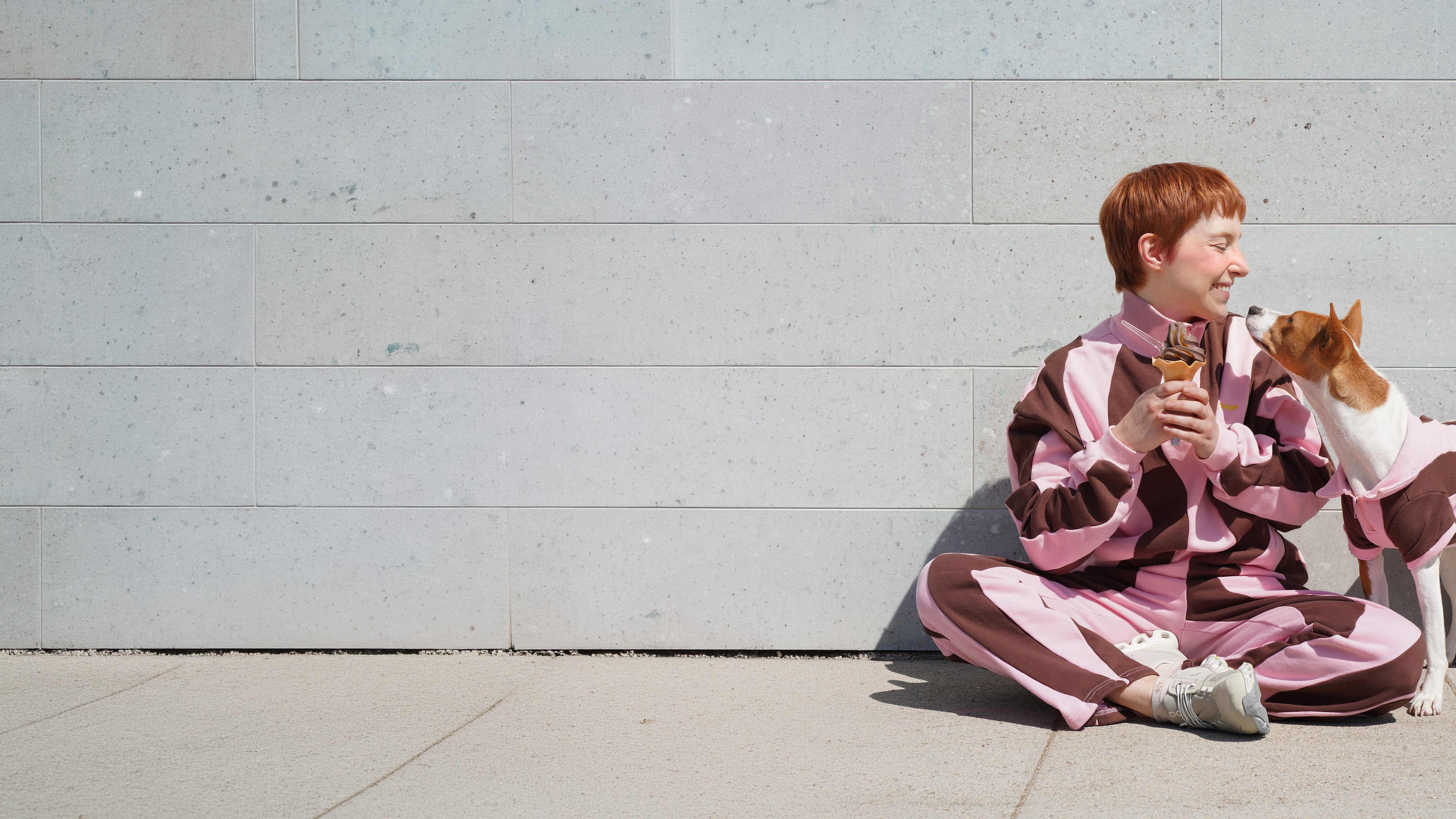 Woman sitting with her dog, both wearing matching pink and brown Sunbean tracksuits, sharing an ice cream cone against a stone wall